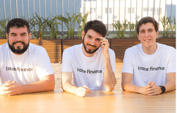 Three smiling male co-founders sitting at a light wooden outdoor table, all wearing matching white t-shirts featuring the "trace finance" company logo.